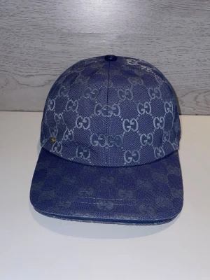 Blå keps från Gucci med GG-mönster - Snygg blå keps från Gucci med klassiskt GG-monogram över hela kepsen. Kepsen är tillverkad i denimliknande material och har justerbar rem baktill. Insidan är fodrad i ljus textil och har märkets etikett. Perfekt för en trendig streetstyle-look.