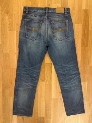 Blå slitna jeans från Nudie Jeans 30/30 - Säljer ett par blå jeans från Nudie Jeans med slitningar och distressed detaljer framtill. Klassisk femficksmodell med raka ben och kontrastsömmar på bakfickorna. Tillverkade i denim, perfekt för en avslappnad och trendig look.