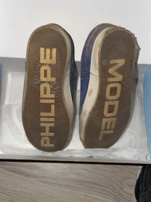 Blå Philippe Model sneakers - Snygga blå sneakers från Philippe Model med vita och bruna detaljer. Skorna har en låg profil, vita snören och en ikonisk logga på sidan. Ovandelen är i mocka och textil, sulan är vit med PHILIPPE MODEL-tryck undertill. Perfekta för en trendig streetwear-look.