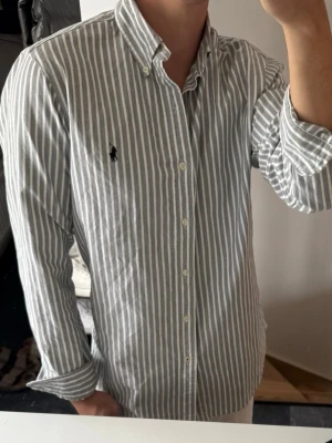 Randig skjorta från Ralph Lauren - Säljer en klassisk vit och grå randig skjorta från Ralph Lauren med button-down krage och broderad logga på bröstet. Skjortan har långa ärmar, normal passform och är tillverkad i mjuk bomull. Perfekt för en clean och stilren look. Lite missfärgad i armhålorna, annars mycket bra skick