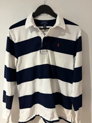 Randig långärmad rugbytröja Polo Ralph Lauren - Klassisk långärmad rugbytröja från Polo Ralph Lauren med breda marinblå och vita ränder. Tröjan har krage, knappar i halsen och röd broderad logga på bröstet. Tillverkad i mjuk bomull för en bekväm känsla.