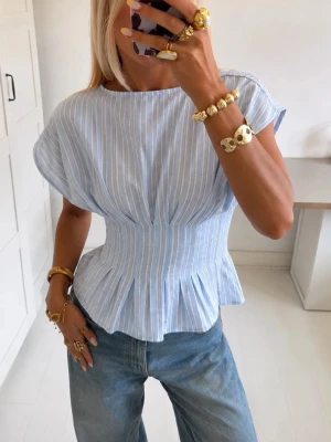Blåvit randig blus med markerad midja - Snygg blåvit randig blus med korta ärmar och markerad midja som ger en feminin siluett. Blusen har lätt volang nedtill och är tillverkad i ett tunt, skönt bomullsmaterial. Perfekt till jeans eller kjol för en fräsch look.