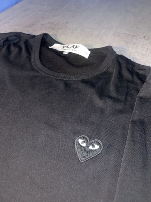 Svart långärmad topp Comme des Garçons - Säljer en svart långärmad topp från Comme des Garçons PLAY med det ikoniska hjärta-märket broderat på bröstet. Toppen har rund halsringning och är gjord i mjuk bomull som känns skön mot huden. Perfekt för dig som gillar stilrena plagg med en twist.