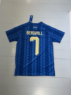 Blå Sverige fotbollströja Adidas | Bergvall  - Säljer en blå Sverige fotbollströja från Adidas med gult tryck, nummer 7 och namnet Bergvall på ryggen. Tröjan har korta ärmar, gul krage och klassiska Adidas-ränder på axlarna. Materialet är lätt och andas, perfekt för träning eller match.