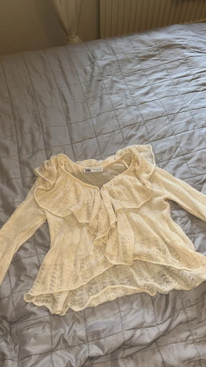 Beige stickad volangblus från Zara - Supersöt beige blus från Zara med volanger längs halsringningen och framtill. Blusen har lång ärm och är stickad i ett lätt, luftigt material med dekorativa hålmönster. Perfekt för dig som gillar romantiska detaljer och en feminin vibe.