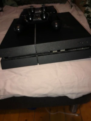 PlayStation 4 med två handkontroller - Säljer en svart PlayStation 4 från Sony tillsammans med två matchande svarta DualShock 4-handkontroller. Konsolen har en stilren, kantig design i matt och blank plast. Perfekt för gamingkvällar och multiplayer med vänner.