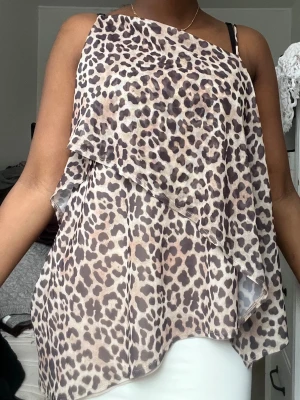 Leopardmönstrad omlott topp - Säljer en luftig och asymmetrisk topp med leopardmönster. Toppen har omlott-design och är ärmlös med ett lager av tunn, transparent polyester. Perfekt för dig som vill sticka ut med ett djuriskt print.