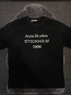 Acne Studios Logo Tee Black - Riktigt fet t-shirt från Acne Studios med det klassiska “Stockholm 1996”-trycket på bröstet 🔥 En av deras mest populära modeller som ger en clean men samtidigt tydlig Acne-look.  Märkt storlek S men är oversized i passformen och passar därför även M utan problem. Fint skick och har mycket kvar att ge.