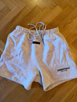 Essentials Fear of God shorts ljusgrå - Säljer ett par ljusgrå shorts från Essentials Fear of God med snörning i midjan och elastisk resår. Shortsen har en avslappnad passform och logga broderad på vänster ben. Perfekta för chill dagar eller streetwear-stil.