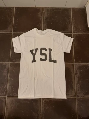 Vit YSL t-shirt med tryck - Snygg vit t-shirt från YSL med stort svart tryck på bröstet. Klassisk rund hals och korta ärmar. Perfekt till jeans eller shorts för en clean och trendig look.