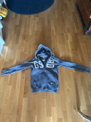 Grå vintage zip hoodie från G-Star RAW - Snygg grå hoodie med dragkedja från G-Star RAW. Stor vit brodyrtext 'GS RD' på bröstet och en patch nertill. Huva med vita snören och känguruficka framtill. Perfekt för en avslappnad vintage streetwear-look.