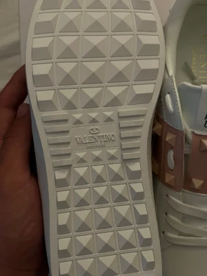 Valentino Garavani vita sneakers - Säljer ett par vita sneakers från Valentino Garavani med rosa detaljer och nitar på sidan. Skorna har en cool sula med rutmönster och klassisk logga under. Materialet är läder och designen är modern och edgy.