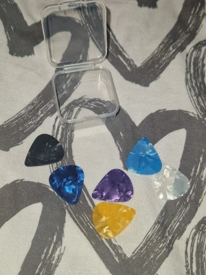 Plektrum - Säljer ett set med sex gitarrplektrum i olika färger: svart, blå, ljusblå, lila, vit och gul. Plektrumen är tillverkade i plast med skimrande yta och kommer med en liten genomskinlig förvaringsask. Helt nya och oanvända.