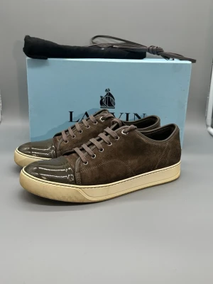 Bruna Lanvin sneakers i mocka - Snygga bruna sneakers från Lanvin med ovandel i mocka och glansig tå i lack. Skorna har beige sula och matchande bruna skosnören. Klassisk låg modell som passar till många olika stilar.