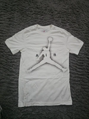 Vit Jordan t-shirt med Air-tryck - Säljer en vit t-shirt från Jordan med det klassiska Air-trycket på framsidan. T-shirten har korta ärmar och är tillverkad i mjuk bomull. Perfekt för dig som gillar sportiga och stilrena plagg med ikonisk design.