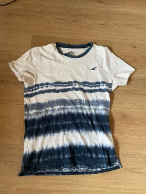 Vit och blå tie-dye t-shirt Hollister - Säljer en snygg vit och blå tie-dye t-shirt från Hollister. T-shirten har korta ärmar, rund halsringning med blå kant och en liten Hollister-logga på bröstet. Tillverkad i mjuk bomull, perfekt för en avslappnad stil.