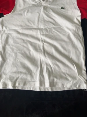 Vit pikétröja från Lacoste - Klassisk vit pikétröja från Lacoste med röda ärmar och den ikoniska gröna krokodilloggan på bröstet. Tröjan har korta ärmar, krage och knappar vid halsen. Perfekt för en sportig och stilren look.