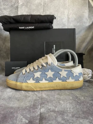 Saint Laurent sneakers med stjärnor - Snygga sneakers från Saint Laurent i ljusblå denim med vita stjärnor längs sidorna. Skorna har vita snören, beige detaljer och en gulaktig sula som ger en vintage vibe. Perfekta för dig som vill sticka ut med cool design och exklusivt märke.