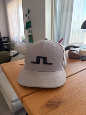 Ljusgrå keps från J.Lindeberg - Snygg ljusgrå keps från J.Lindeberg med svart broderad logga framtill. Klassisk modell med böjd skärm och justerbar baksida. Tillverkad i ett slitstarkt tyg som passar perfekt till streetwear eller casual outfits.  Size 7 alltså 55,8 cm