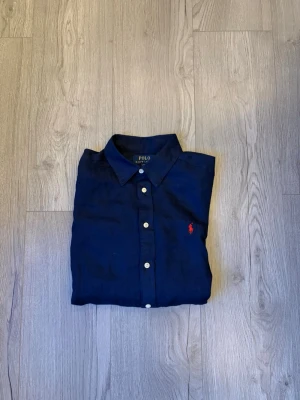 Mörkblå linneskjorta från Polo Ralph Lauren - Klassisk mörkblå skjorta i linne från Polo Ralph Lauren med vit knappstängning och ikonisk röd logga broderad på bröstet. Skjortan har lång ärm och normal passform, perfekt till en stilren look. Skjortan är använd få gånger och har inga defekter! Skjortan är L i barnstorlek men jag skulle säga att den passar en XS. Skriv vid minsta fundering🤩✅