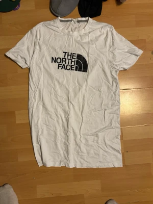 Vit t-shirt från The North Face - Säljer en vit t-shirt från The North Face med klassisk svart logga på bröstet. T-shirten har rund halsringning och korta ärmar. Tillverkad i mjuk bomull som är skön att bära. Perfekt till en avslappnad stil.