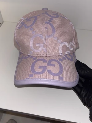 Ljuslila keps från gg - Säljer en snygg ljuslila keps från Gg med det klassiska GG-mönstret i lila över hela kepsen. Kepsen har böjd skärm och är tillverkad i ett strukturerat tyg med matchande lila kant runt skärmen. Perfekt för dig som vill sticka ut med en trendig.