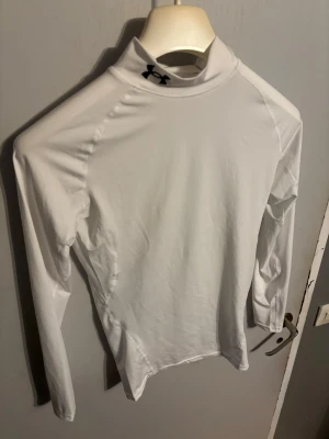 Vit kompressionströja Under Armour - Säljer en vit långärmad kompressionströja från Under Armour med HeatGear-teknologi. Tröjan har hög krage och är tillverkad i ett stretchigt funktionsmaterial som transporterar bort fukt. Perfekt för träning eller sportaktiviteter.