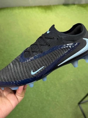 Nike svart/blå fotbollssko - Snygg fotbollssko från Nike i svart med blå detaljer och ljusblå Swoosh-logga. Skon har en strumpliknande passform, snörning och mönstrad ovandel i syntetmaterial för bra bollkontroll. Sulan har fasta dobbar i blått för spel på gräs.