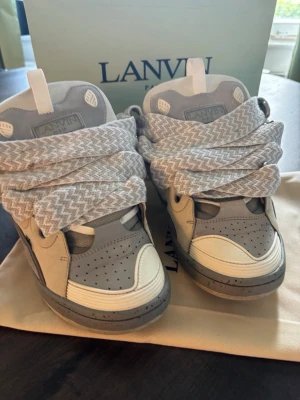 Lanvin gråvita sneakers med breda snören - Lanvin sneakers i grått och vitt med breda, mönstrade snören. Skorna har en chunky siluett, perforerad tåbox och detaljer i både läder och mocka. Märkeslogga på plösen och cool retrovibe. Perfekta för dig som gillar statement sneakers.