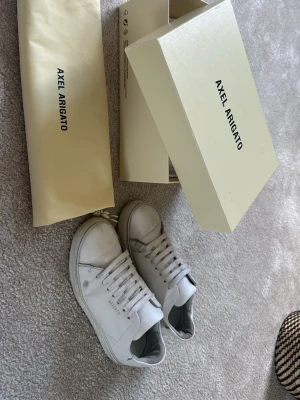 Vita sneakers från Axel Arigato - Snygga vita sneakers från Axel Arigato i minimalistisk design. Skorna är låga med klassisk siluett, vita skosnören och diskret logga på sidan. Tillverkade i läder för en clean och stilren look. Perfekta för dig som gillar enkel och modern stil.