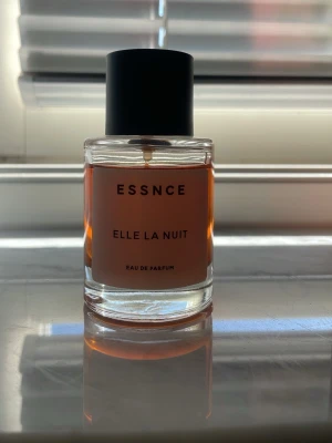 Essnce Elle La Nuit Eau de Parfum - En stilren parfymflaska från Essnce med doften Elle La Nuit. Flaskan är cylindrisk i klart glas med svart lock och innehåller en bärnstensfärgad vätska. Minimalistisk etikett med svart text ger en modern känsla. Perfekt för dig som gillar eleganta och trendiga dofter.