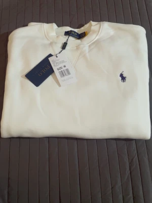 Vit sweatshirt från Polo Ralph Lauren - Klassisk vit sweatshirt från Polo Ralph Lauren med rund halsringning och broderad marinblå logga på bröstet. Tröjan har ribbade muddar vid ärmslut och nederkant. Perfekt för en clean och stilren look.