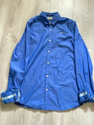 Blå skjorta från Scotch & Soda - Snygg blå skjorta från Scotch & Soda med klassisk krage och vita knappar. Skjortan har en bröstficka och detaljer på insidan av manschetterna. Tillverkad i mjuk bomull, perfekt för en stilren look.