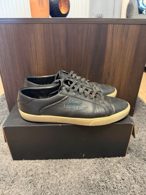 Saint Laurent sneakers - Svarta sneakers från Saint Laurent i mjukt läder med blå logga på sidan. Skorna har svarta skosnören och en beige sula som ger en snygg kontrast. Klassisk låg modell som passar till många olika stilar.