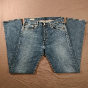 Levi's 501 blå jeans - Klassiska blå Levi's 501 jeans med raka ben och femficksdesign. Byxorna har knappgylf, tydliga kontrastsömmar och den ikoniska läderpatchen bak i midjan. Perfekta för en avslappnad och tidlös stil.