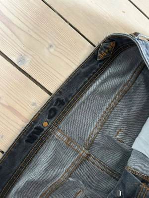 Mörkgrå jeans från NN07 - Säljer ett par mörkgrå jeans med snygg tvättad effekt och klassisk femficksmodell. Jeansen har orange kontrastsömmar och rak passform. Perfekta för dig som gillar en avslappnad men ändå stilren vibe.
