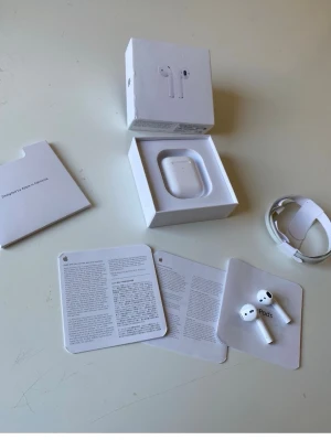 Apple AirPods trådlösa hörlurar - Säljer ett par vita Apple AirPods med laddningsetui och originalförpackning. Hörlurarna är trådlösa och har en stilren, minimalistisk design i plast. Medföljer gör laddningskabel och manualer. Perfekta för musik, samtal och vardagsbruk.
