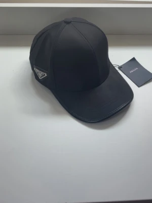 Svart keps från Prada i nylon - Snygg svart keps från Prada med klassisk böjd skärm och ikonisk metallogo på sidan. Tillverkad i glansigt nylonmaterial som ger en modern och clean look. Perfekt accessoar för dig som gillar exklusiva streetwear-vibbar.