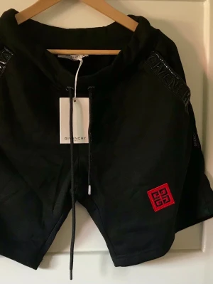 Svarta shorts från Givenchy - Svarta shorts från Givenchy med snörning i midjan och en röd Givenchy-logga på vänster ben. Shortsen har detaljer med Givenchy-tryck på sidorna och är tillverkade i ett mjukt material som känns lyxigt. Perfekta för en trendig och avslappnad stil.