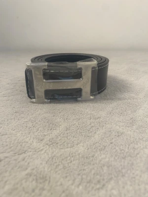 Svart skärp med H-spänne - Snyggt svart skärp  med ett stort, silverfärgat H-spänne. Skärpet har vita kontrastsömmar längs kanterna och är gjort i slätt skinn. Perfekt accessoar för att lyfta vilken outfit som helst.