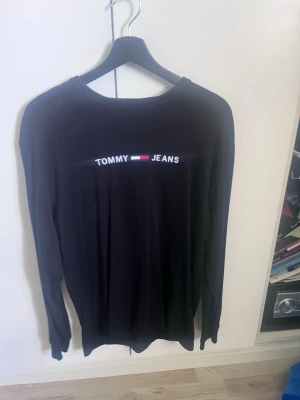 Svart långärmad T-shirt Tommy Jeans - Svart långärmad topp från Tommy Jeans med rund halsringning och logga framtill i vitt, rött och blått. Enkel och stilren design som passar till det mesta. Tillverkad i mjukt material för en bekväm känsla.