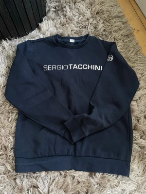 Mörkblå sweatshirt Sergio Tacchini - Har små fläckar som inte syns långt ifrån . Man ser de på 2 bilden Mörkblå sweatshirt från Sergio Tacchini med vit logotyp och text framtill. Klassisk rund halsringning och ribbade muddar vid ärmslut och nederkant. Perfekt för en avslappnad och sportig stil.