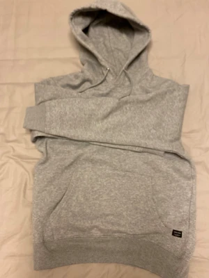Grå hoodie från Jack & Jones - Klassisk grå hoodie från Jack & Jones med huva och dragsko. Tillverkad i mjukt sweatshirtmaterial och har en stor magficka framtill. Perfekt för en avslappnad och stilren look.
