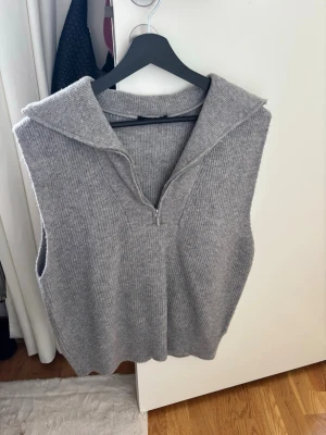 Grå stickad väst från Gina Tricot - Mysig grå stickad väst från Gina Tricot med krage och half zip-dragkedja framtill. Perfekt att slänga över en skjorta eller t-shirt för en chill och trendig look. Västen har en ribbad struktur och är i en neutral grå färg som passar till allt.
