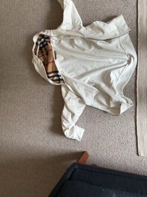 Vit hoodiejacka från Burberry - Säljer en vit hoodiejacka från Burberry med klassiskt rutigt foder i huvan. Jackan har dragkedja framtill och en avslappnad passform. Perfekt för dig som vill ha en stilren och tidlös look med en touch av lyx.
