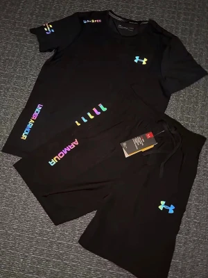 Svart träningsset från Under Armour - Säljer ett svart träningsset från Under Armour med t-shirt och shorts. Båda plaggen har färgglada loggor och textdetaljer i regnbågsfärger. Perfekt för gymmet eller löpning. Materialet är lätt och andas, vilket gör det bekvämt under träning. Storlek S, helt sprillans ny 