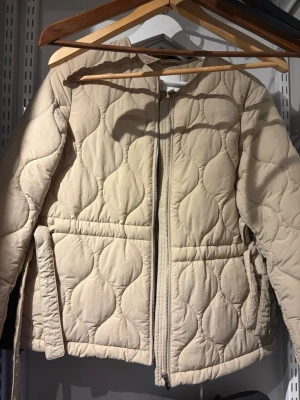 Beige quiltad pufferjacka - Snygg beige pufferjacka med quiltat mönster och dragkedja framtill. Jackan har två fickor med lock och en rak passform. Perfekt för kalla dagar och ger en trendig look. Materialet känns mjukt och isolerande.