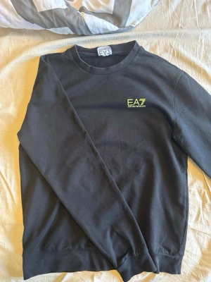 Svart stickad tröja från EA7 Armani - Snygg svart stickad tröja från EA7 Emporio Armani med rund halsringning och diskret EA7-logga i gult på bröstet. Tröjan har långa ärmar och ribbade muddar vid ärmslut och nederkant. Perfekt för en stilren och avslappnad look.