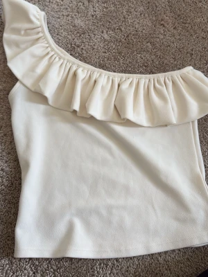 Vit axelbandslös topp med volang - Snygg vit axelbandslös topp med bred volang längs ena axeln. Perfekt för dig som vill ha en trendig och stilren look. Toppen har en mjuk och lätt struktur som känns skön mot huden.