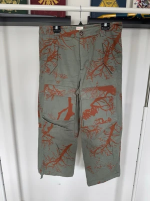 Gröna cargobyxor med orange mönster - Unika cargobyxor i grönt med orange grenmönster. Byxorna har flera stora fickor framtill och en bakficka, samt bred passform och raka ben. 